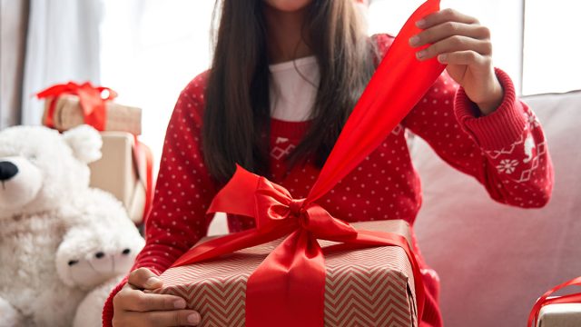 Smiling kid girl holding red ribbon in hands opening Christmas gift box at home, close up view. Happy cute child wearing santa hat receiving xmas New Year present sitting on couch celebrating holiday.