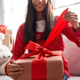 Smiling kid girl holding red ribbon in hands opening Christmas gift box at home, close up view. Happy cute child wearing santa hat receiving xmas New Year present sitting on couch celebrating holiday.