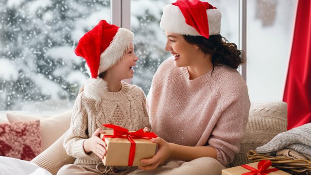 Merry Christmas and Happy Holidays. Cheerful mom and her cute daughter girl exchanging gifts. Parent and little child having fun near tree indoors. Morning Xmas.