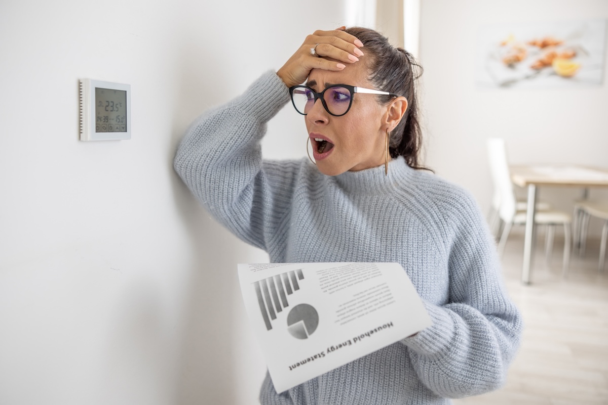 upset woman holding a heating bill and looking at her thermostat