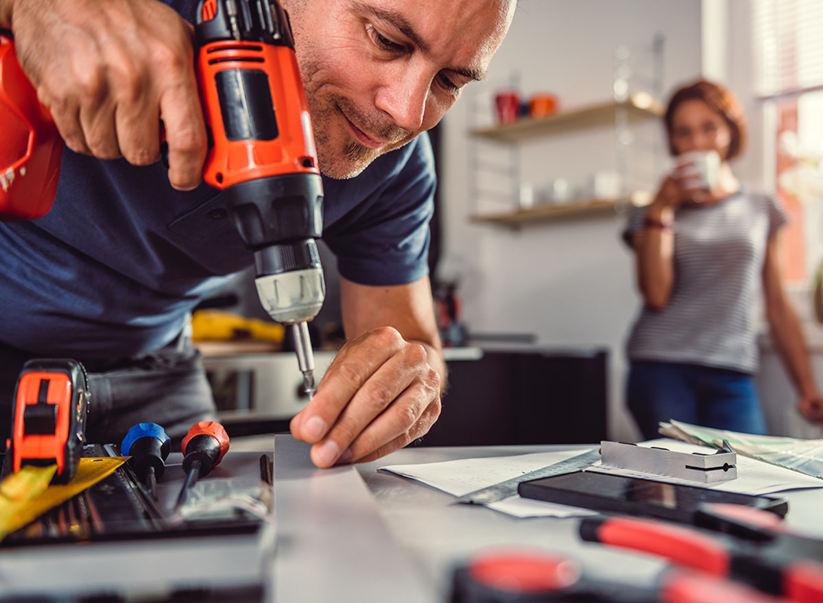 Men renovating kitchen and using cordless drill in the background standing his wife and drinking coffee