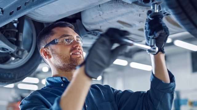 Portrait Shot of a Handsome Mechanic Working on a Vehicle in a Car Service. Professional Repairman is Wearing Gloves and Using a Ratchet Underneath the Car. Modern Clean Workshop.