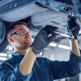 Portrait Shot of a Handsome Mechanic Working on a Vehicle in a Car Service. Professional Repairman is Wearing Gloves and Using a Ratchet Underneath the Car. Modern Clean Workshop.