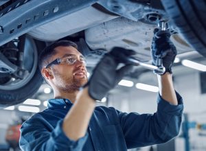 Portrait Shot of a Handsome Mechanic Working on a Vehicle in a Car Service. Professional Repairman is Wearing Gloves and Using a Ratchet Underneath the Car. Modern Clean Workshop.