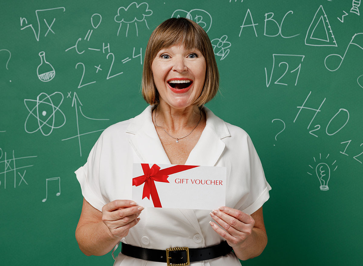 Fun teacher mature elderly senior woman 55 wear white shirt hold gift certificate coupon voucher card for store isolated on green wall chalk blackboard background studio. Education in school concept