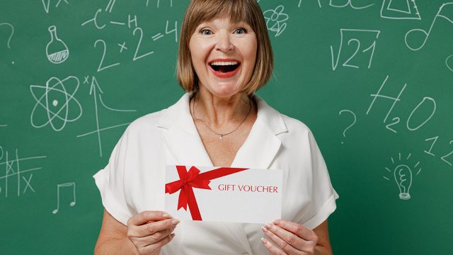 Fun teacher mature elderly senior woman 55 wear white shirt hold gift certificate coupon voucher card for store isolated on green wall chalk blackboard background studio. Education in school concept