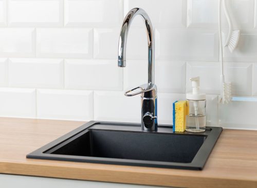 An empty black kitchen sink in a kitchen with a chrome faucet, wooden countertop, and white subway tile backsplash with dishwashing liquid, brush and sponge, banner with copy space