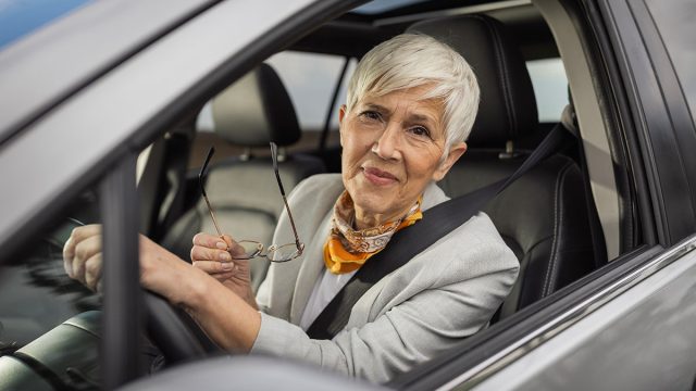 Senior woman with short silver hair smiles while holding her glasses in car. She is wearing stylish outfit and appears content during her drive