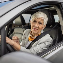Senior woman with short silver hair smiles while holding her glasses in car. She is wearing stylish outfit and appears content during her drive