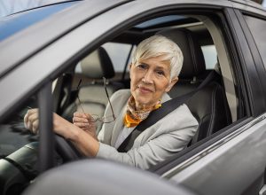 Senior woman with short silver hair smiles while holding her glasses in car. She is wearing stylish outfit and appears content during her drive