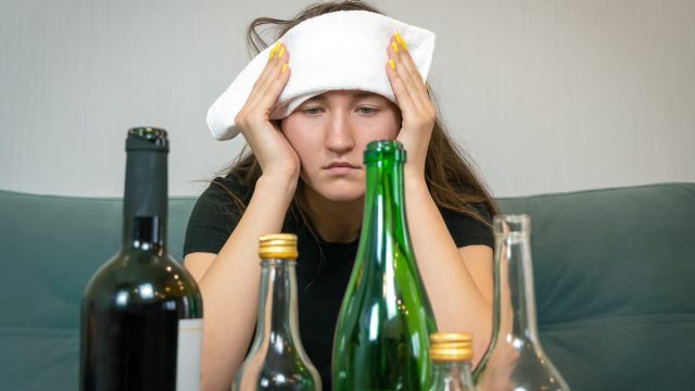 Young woman with a white towel on her forehead from a headache sits in front of empty bottles of alcohol. Concept of Monday morning, morning after drinking alcohol