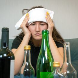 Young woman with a white towel on her forehead from a headache sits in front of empty bottles of alcohol. Concept of Monday morning, morning after drinking alcohol