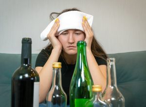 Young woman with a white towel on her forehead from a headache sits in front of empty bottles of alcohol. Concept of Monday morning, morning after drinking alcohol