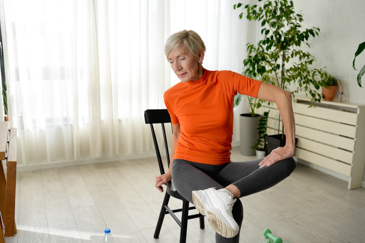 mature woman in an orange top stretching her hips while sitting on a chair