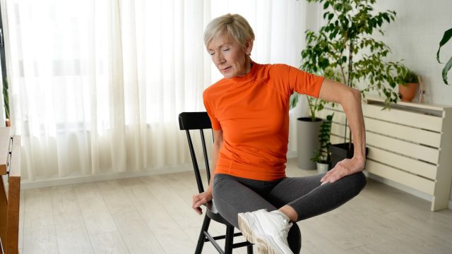 mature woman in an orange top stretching her hips while sitting on a chair
