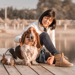 Portrait of beautiful young woman playing with dog by the river. Happy woman sitting on the wooden pier with her dog Basset Hound. Woman with puppy.