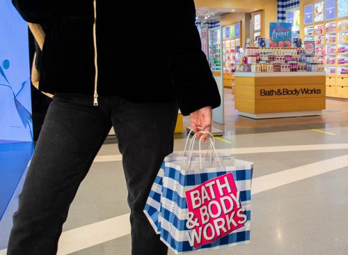 A female customer holds in her hand paper bags branded "Bath and body works" in a blue and white check and a pink logo on the background of the entrance to the store