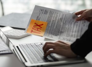A person handles tax forms while using a laptop, highlighting the critical task of preparing for tax season.