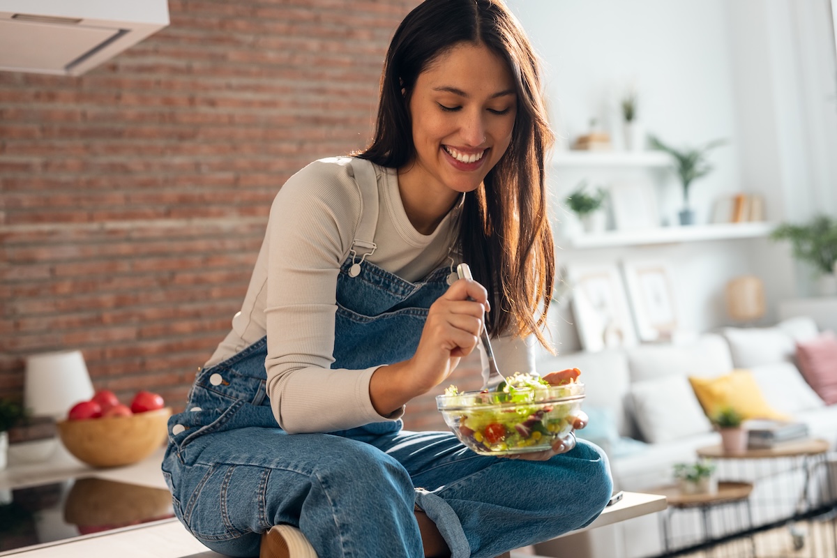 smiling woman sitting on kitchen counter eating a salad