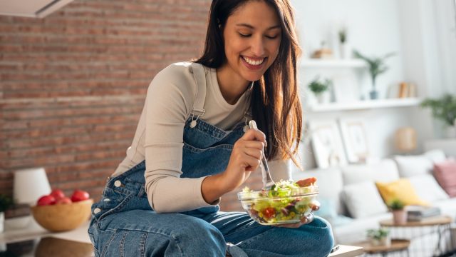 smiling woman sitting on kitchen counter eating a salad