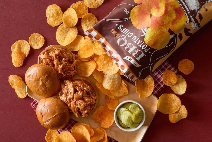 Trader Joe's BBQ Flavored Potato Chips shown on deep red surface; a tray with 2 pulled chicken sandwiches and a cup of Kosher Dill Pickle Chips