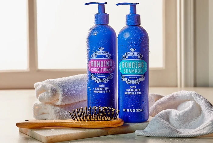 Trader Joe's Bonding Shampoo and Conditioner shown in a bright white surface and windown background, white towels and a hairbrush