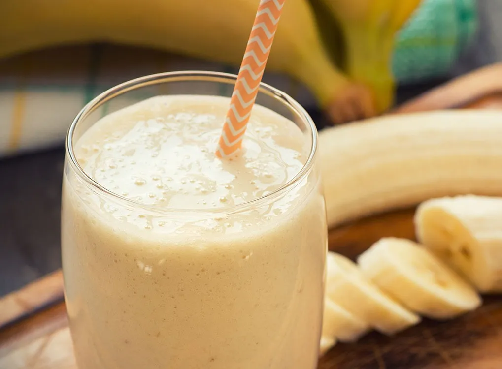 close up of a banana smoothie in a glass with a straw on a wooden cutting board with bananas in the background
